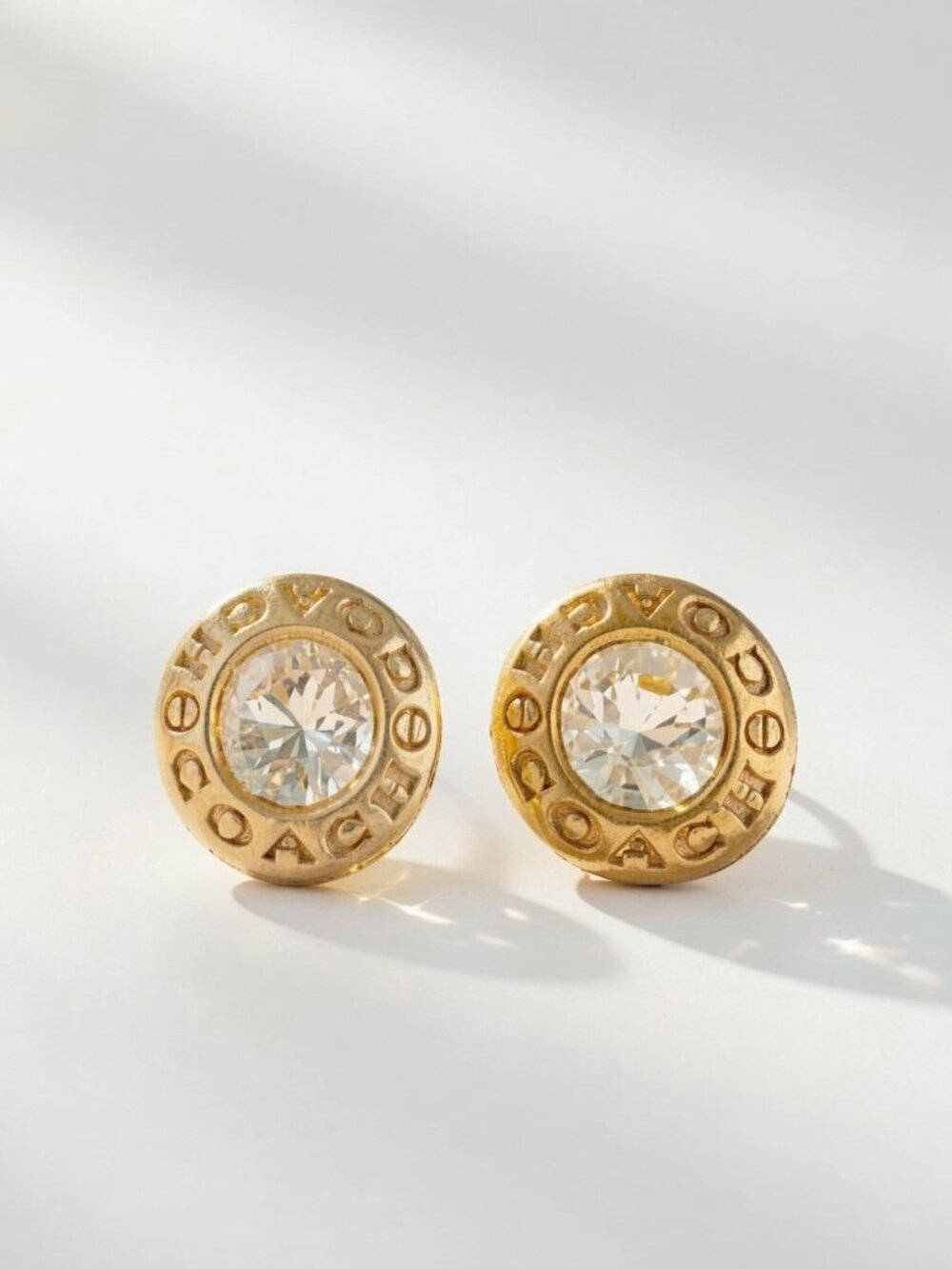Coach gold earrings with clear crystal center and 'COACH' lettering around the e